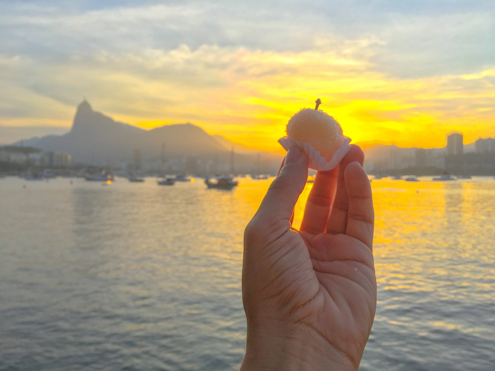 A classic Brazilian beijinho: coconut candy ball in a paper cup with a clove on top, held against a Rio sunset