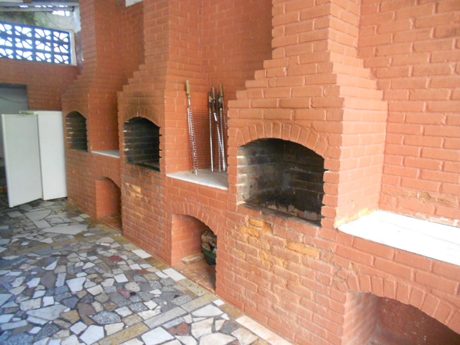 Three built-in brick churrasqueiras with chimneys, long metal skewers, and a grate leaning against the brick