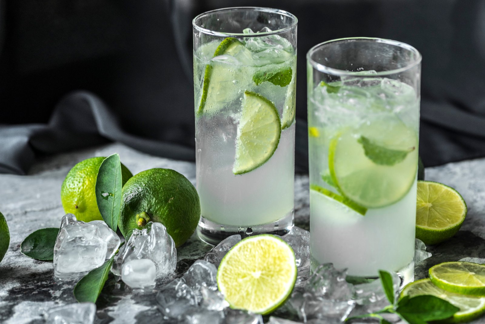Two caipirinhas in tall glasses with lime wedges and ice