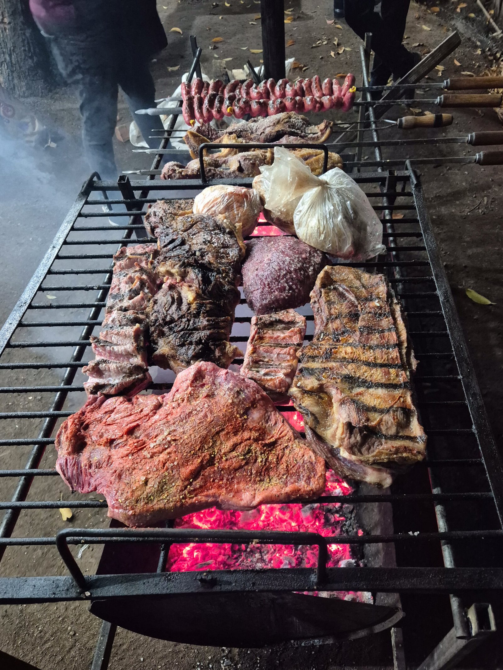 Meat on a grate in the foreground and long wooden-handled skewers of sausage and beef on a rack above a charcoal fire