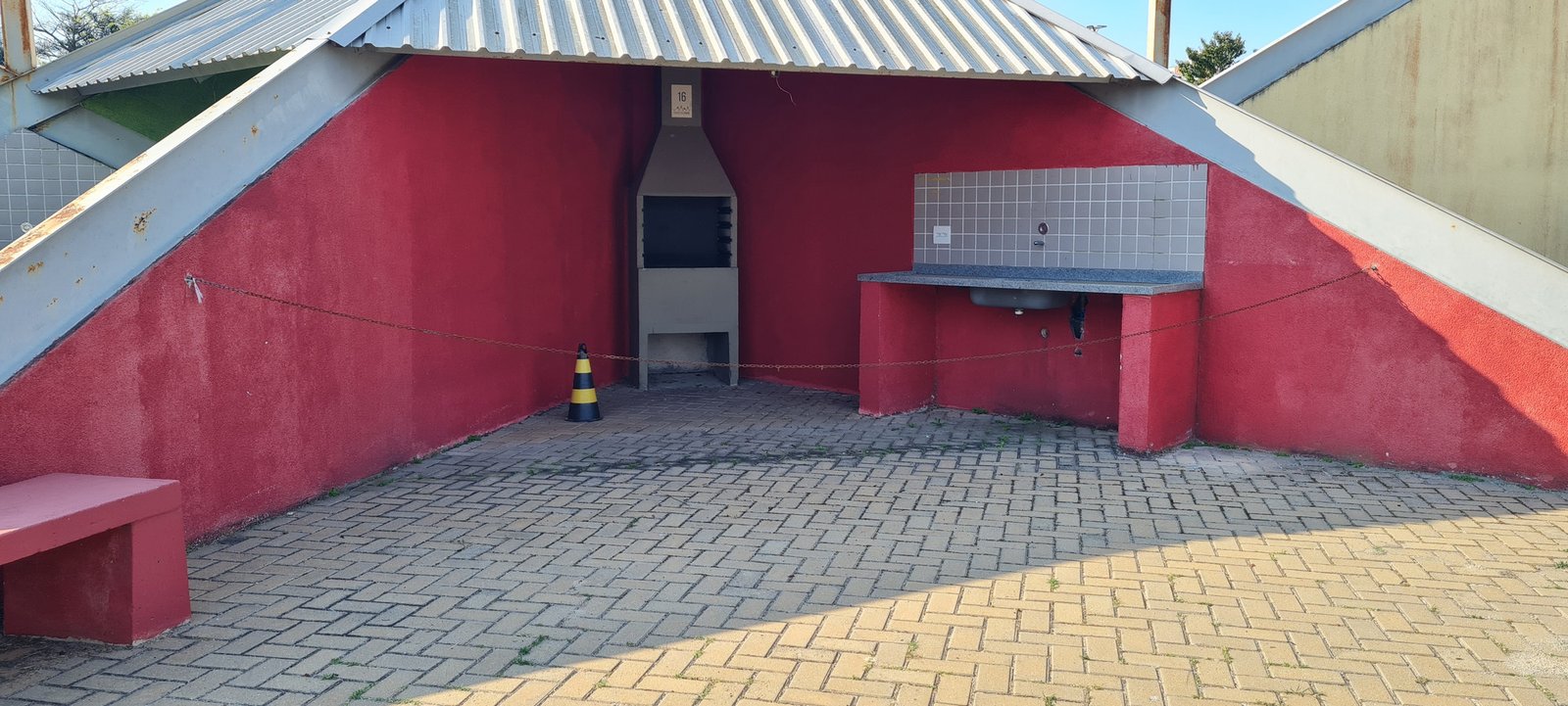 Standalone gray precast concrete churrasqueira with chimney, firebox, and lower storage, in a red-walled park alcove