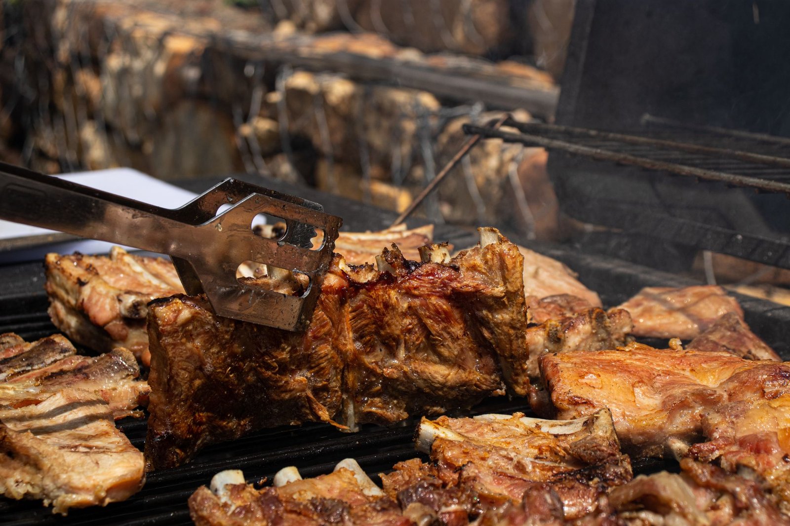 Pork ribs grilling on a charcoal grate, bones visible, lifted with tongs
