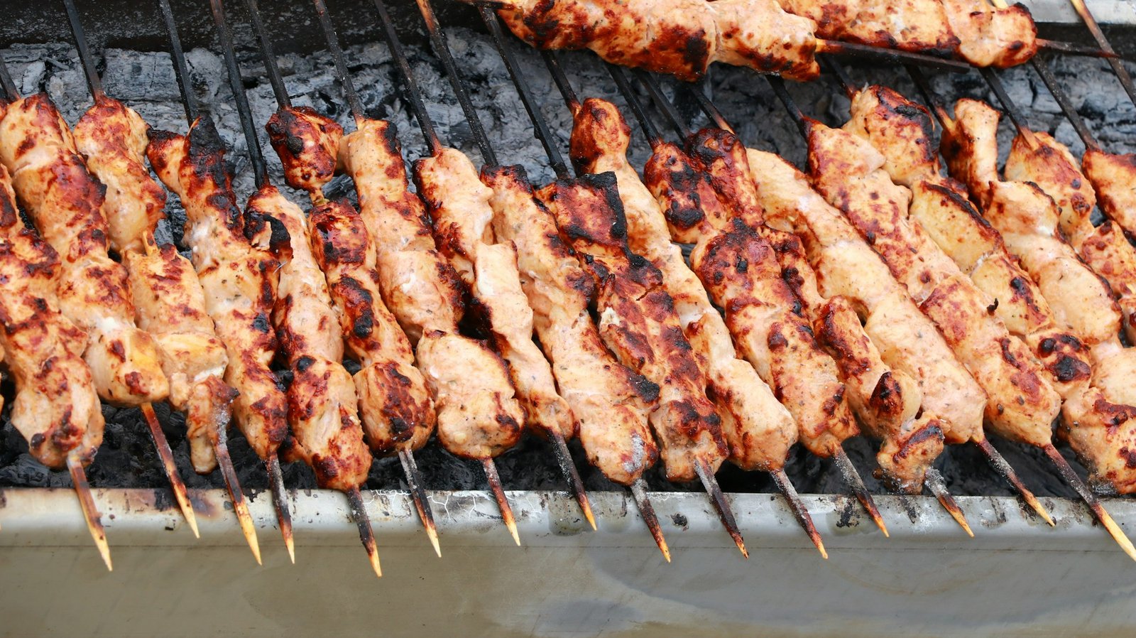 Many chicken cubes on wooden skewers cooking over a bed of charcoal embers
