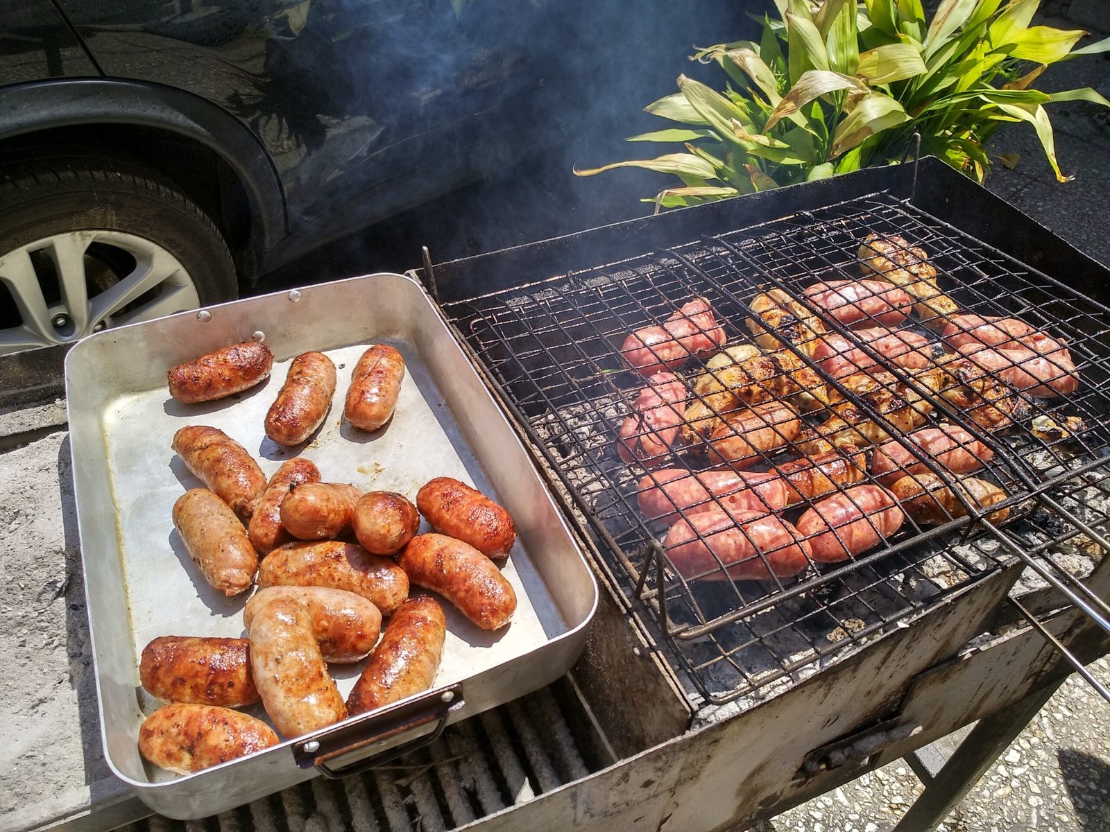 Fresh pork sausages grilling over charcoal, with already-browned links in a pan